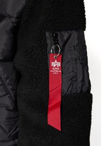 Veste noire texturée avec poche en nylon noir et étiquette rouge en tissu pour porte-clés indiquant Alpha Industries Inc. U.S.A., suspendue à une tirette de fermeture éclair.