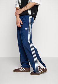 Marinfärgade träningsbyxor med vita sidostreck, med en avslappnad passform och dragkedjeslitsar vid anklarna. Adidas-logotypen på vänster lår.