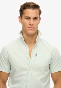 Camicia verde chiaro a maniche corte realizzata in morbido cotone, con colletto button-down e taschino sul lato sinistro con un piccolo dettaglio del logo.