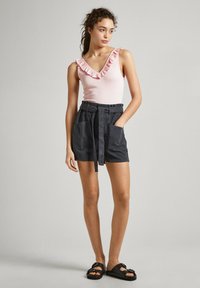 Top senza maniche di un delicato rosa pallido con scollo a volant, abbinato a pantaloni corti scuri con cintura e tasche, indossato con sandali neri.