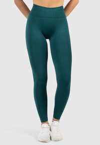 Türkise high-waisted Leggings aus glattem, dehnbarem Material, mit einem enganliegenden Design, das bis zu den Knöcheln reicht, kombiniert mit weißen Sportschuhen.
