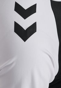 Zwart dubbel-chevronlogo op wit textiel, close-up van sportkledingmateriaal met een zwart zijpaneel.