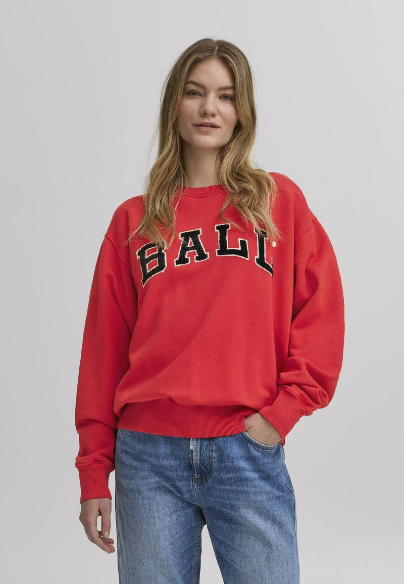Rød hettegenser med "BALI" i svarte bokstaver på brystet, har en avslappet passform, ribbede mansjetter og myk stoff, kombinert med blå jeans.