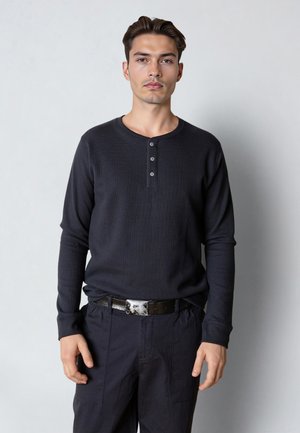 Jeune homme aux cheveux courts et foncés, portant un t-shirt henley à manches longues noir et un pantalon noir avec une ceinture à boucle argentée, debout devant un fond uni.
