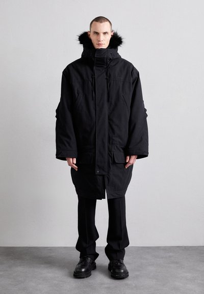 Zwarte geïsoleerde parka met een met bont gevoerde capuchon, voorzien van een ritssluiting aan de voorkant, grote zakken en een losvallend ontwerp. Gecombineerd met zwarte broeken en laarzen.