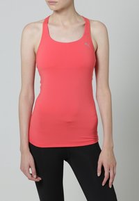 Nainen, joka on pukeutunut korallinväriseen Puma ilmanhihaiseen urheilutoppia ja mustiin legginsseihin, seisoo yksinkertaista vaaleanharmaata taustaa vasten.