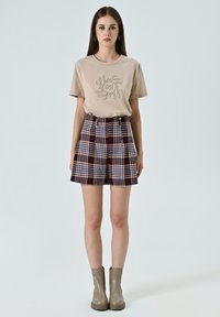 T-shirt beige con grafica testurizzata, abbinata a pantaloni corti a scacchi marroni e bianchi. Il modello indossa stivaletti beige, in piedi su uno sfondo neutro.