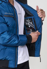 Plava pufasta jakna s crnim akcentima, koja ima zippered džep na prsima, rebraste manžetne i glatku unutrašnjost s informativnom oznakom.