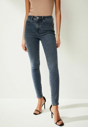 Smala jeans i mörkblå denim, hög midja med fem fickor, kontrasterande sömmar och knappstängning fram. Bärs med svarta remsandaler.