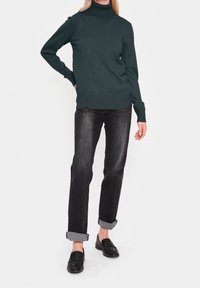 Pull à col roulé vert avec poignets côtelés, associé à un jean denim foncé retroussé au bas et des mocassins noirs. Texture en maille lisse.