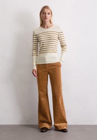 Vrouw die tegen een witte muur staat, gekleed in een lange mouwshirt met beige en bruine strepen, een bruine wijde broek en bruine schoenen.