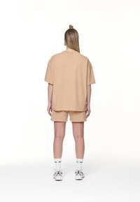 Donna in piedi con le spalle rivolte verso di noi, indossa una maglietta oversize beige e pantaloni corti, calze bianche e sneakers bianche, su uno sfondo bianco semplice.