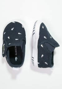 Námořnické plátěné nazouvací boty s bílým logem Lacoste a malými bílými logy potištěnými po celém povrchu. Bílé gumové podrážky s černými pruhy.