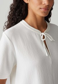 Blouse blanche avec un tissu texturé, à manches courtes, et une ouverture en forme de trou de serrure au niveau du cou, dotée d'une fermeture à nouer faite du même matériau.
