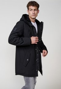 Melnā parka jaka ar kapuci, ar priekšējo rāvējspodu, sprādzēm un priekšējiem kabatām. Tekstūrēta auduma ar elegantu, minimālistisku dizainu.