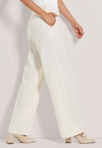 Witte gestreepte wijde broek met hoge taille, voorzien van zijzakken. Draagt een paar beige blockhakken. Glad stoftextuur.