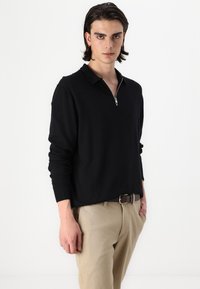 Schwarzes Langarmpolo mit Reißverschlusskragen, aus weichem Stoff, kombiniert mit beigen Hosen und einem braunen Gürtel.