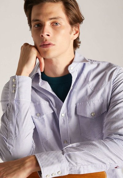 Gestreept button-up shirt in paars en wit. Heeft twee borstzakken, drukknoopsluiting en een gestructureerde stof. Draagt over een groene onderhemd.