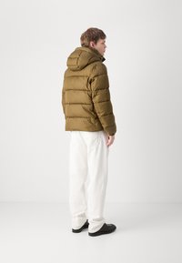 Olivine žalsvas pūkinis džemperis su gobtuvu, su quilted tekstūra ir užtrauktuku, dėvimas su baltais plačiais keliais ir juodais batais.