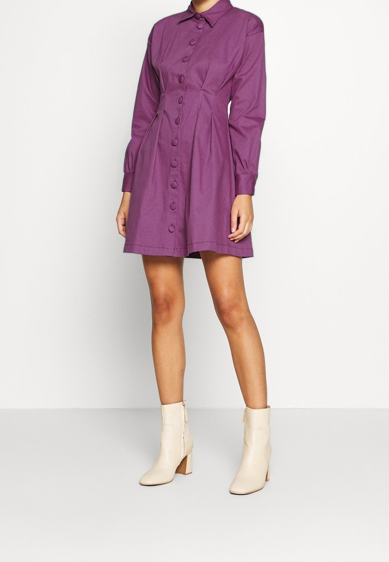 Femme portant une robe violette boutonnée à manches longues, associée à des bottines beige clair, debout devant un fond blanc uni.