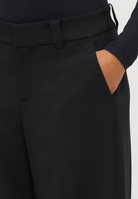 Melnas bikses no gludas auduma, ar plakanu priekšpusi un sānu kabatām; ir šaurs jostasviets ar jostas cilpu detaļu.