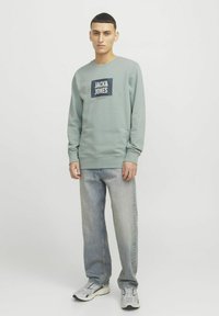 Sudadera verde con un logo cuadrado oscuro en el centro, combinada con jeans azul claro y zapatillas deportivas grises. Diseño simple y casual.
