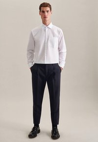 Weißes Hemd aus strukturiertem Stoff mit einer Brusttasche, kombiniert mit dunklen, zulaufenden Hosen und schwarzen Schnürschuhen.