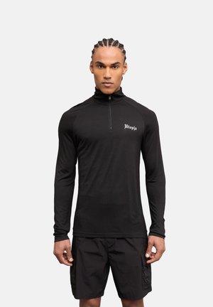 Hombre con trenzas, con una camiseta negra de manga larga con cremallera y el texto "Utopia" y pantalones cortos cargo negros, de pie delante de un fondo liso.