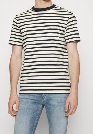 Mand iført en sort og hvid vandretstribet t-shirt med korte ærmer og lyseblå jeans mod en ensfarvet baggrund.