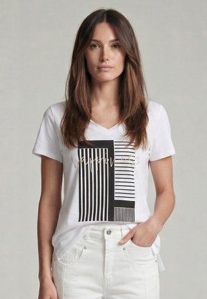 Young woman with long brown hair wearing a white V-neck T-shirt with black and white vertical striped graphic and white pants.
