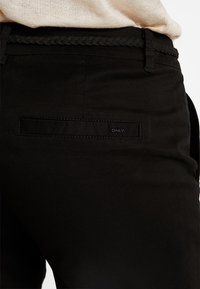 Pantalon en tissu noir avec une taille tressée et une poche arrière ornée d'une étiquette discrète. La texture semble lisse et confectionnée sur mesure.