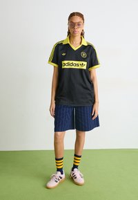 Melnā Adidas kreklā ar dzelteniem apdariem un logo, svītrainā dizaina, apvienots ar jūras zilās svītrainām šortiem, daudzkrāsainām svītrainām zeķēm un rozā kedām.