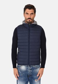 Gilet imbottito blu navy con design a cuciture orizzontali, colletto alto e zip frontale; presenta un hoodie grigio sottostante e tasche laterali.
