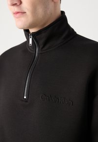 Pulover negru din fleece cu guler înalt, închidere cu fermoar și logo-ul Calvin Klein embosat pe piept. Textură netedă și design minimalist.