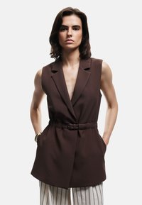 Blazer marrone senza maniche con scollatura a V profonda, spalle strutturate, vita con cintura e tasche laterali, abbinato a pantaloni a gamba larga a righe.