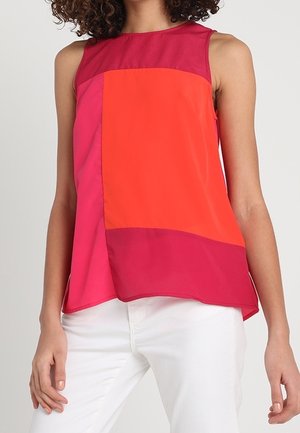 Femme portant un haut sans manches à blocs de couleurs dans des tons rouges et orange avec un pantalon blanc, debout devant un fond uni.