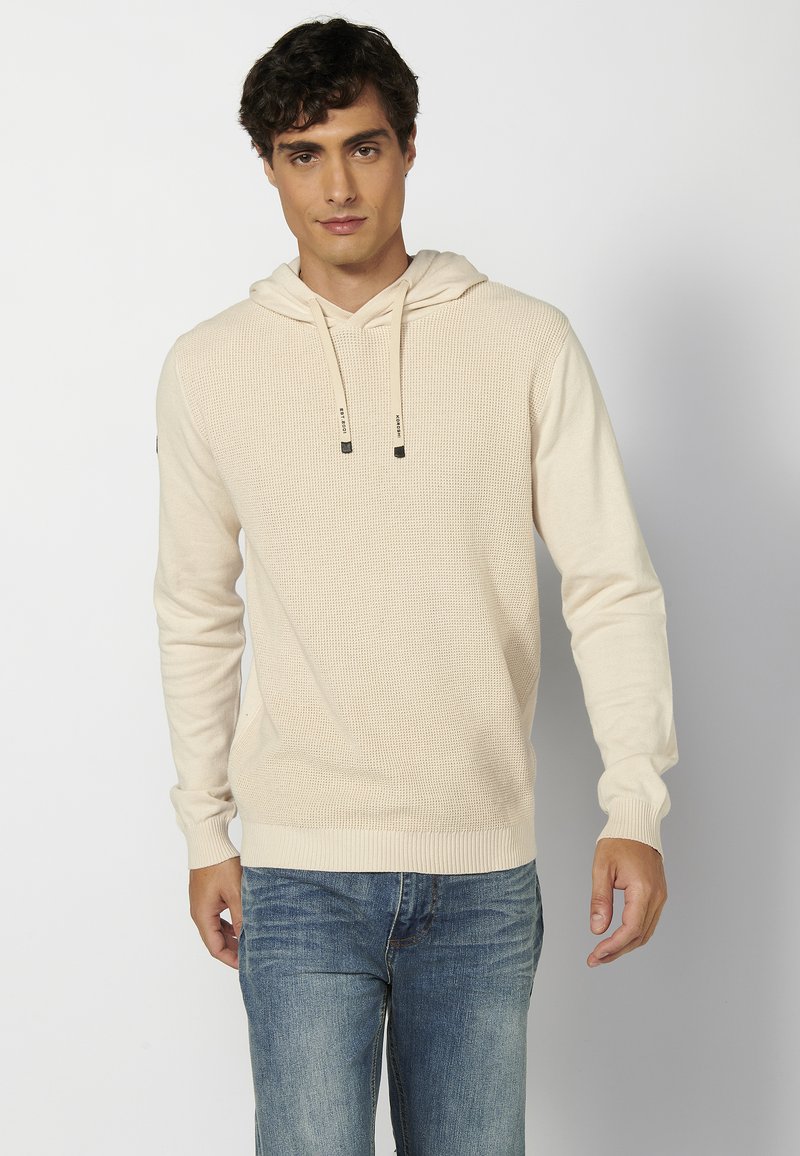 Haut à capuche beige texturé avec un motif côtelé, un col à cordon de serrage et des poignets ajustés, associé à un jean bleu. Style simple et décontracté.