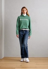 Felpa verde in cotone con logo bianco "Marc O'Polo", abbinata a pantaloni in denim blu e scarpe da ginnastica bianche, con un dolcevita bianco sotto.