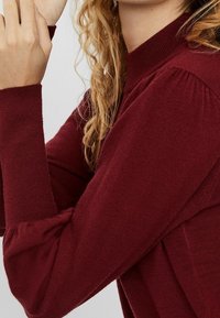 Femme avec des cheveux bouclés portant un pull bordeaux à col montant et à manches longues avec des détails de manches froncées sur un fond clair.