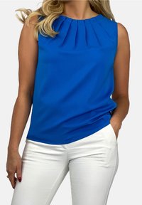 Blå ärmlös topp med plisséad halsringning, gjord av mjukt tyg; kombinerad med vita byxor som har sidofickor och smal passform.