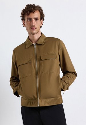 Mann mit lockigem Haar, der eine beige Reißverschlussjacke mit zwei Brusttaschen trägt und mit den Händen in den Taschen vor einem einfarbigen Hintergrund steht.