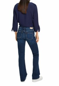 Haut bleu marine avec manches nouées, associé à un jean évasé bleu foncé avec détails de poche et une étiquette beige clair à la taille.