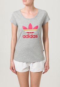 Grå bomullströja med korta ärmar, med en stor rosa och vit Adidas-logotyp och bladdesign över bröstet. Vita shorts.