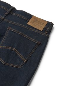 Los pantalones vaqueros de mezclilla oscura presentan costuras naranjas contrastantes, una etiqueta de cuero de marca y un único bolsillo trasero con una textura sutil.