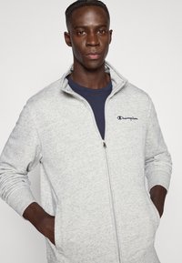 Ανοιχτό γκρι φούτερ με φερμουάρ, κατασκευασμένο από απαλό ύφασμα, με ψηλό γιακά, ασημένιο φερμουάρ και κεντημένο ναυτικό λογότυπο στο στήθος.