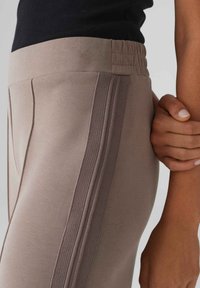 Beige sportbroek met elastische tailleband, voorzien van zijstrepen en een gladde textuur. Close-upweergave die de pasvorm en ontwerpelementen benadrukt.