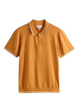 Mann trägt ein senfgelbes kurzärmeliges Poloshirt mit weißem Besatz, steht mit einer Hand in der Tasche vor einer beigen Wand.