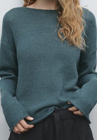 Sininen neulepusero leveällä pyöreällä kaula-aukolla, väljä mitoitus ja pidennetyt hihat, yhdistettynä mustiin korkeavyötäröisiin housuihin.