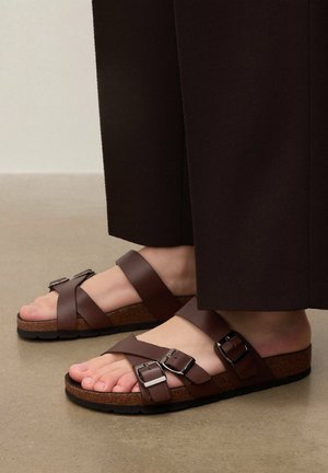 Barna bőrszandált viselő lábak, három állítható csattal és parafa talppal, sötétbarna, széles szárú nadrággal párosítva.