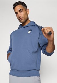 Sweatshirt azul com um bolso frontal, design de golfe bordado no peito, punhos e bainha ribados. Modelo segura um taco de golfe sobre o ombro.